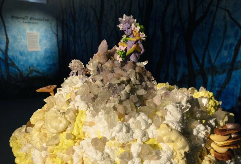 Detail photo of miniature fairy sculptures by Tammy Salzyl. Two fairy figures, one purple and one orange, embrace on top of a hill of crystals, mushrooms and foam. Crystals grow from the purple fairy's head, flowers grow from the orange fairy's head.