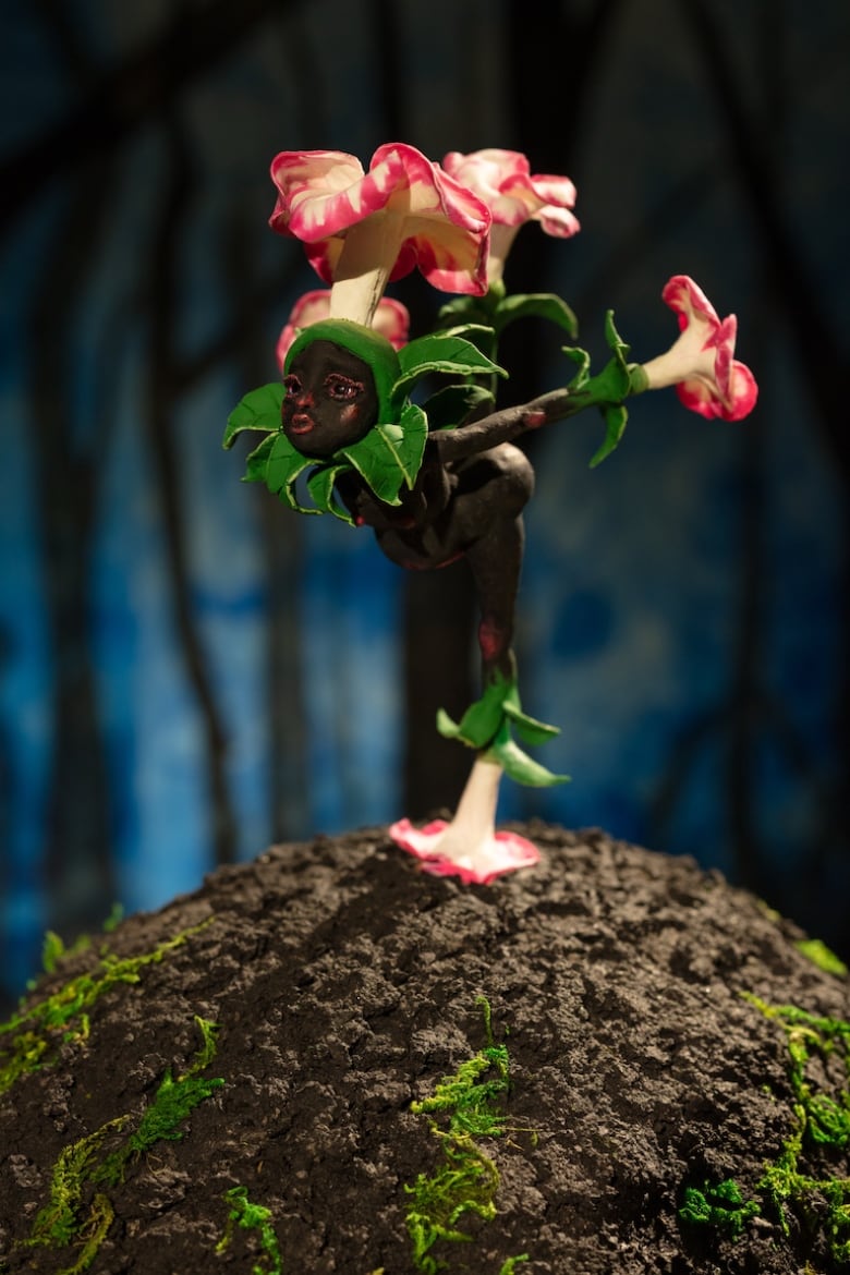 A tiny sculpture of a nude brown-skinned fairy with pink and white flowers for hands and feet, poses like a dancer on a hill of soil.