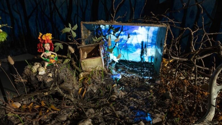 Photo of a diorama by Tammy Salzyl. Mossy ground covers a tabletop. A clay figure of a tiny nude fairy with fiery flower petals on its head kneels at left in a bird's nest, holding a broken speckled egg in its hands. A wooden crate at centre is illuminated to reveal a blue-sky landscape. Gnarled brown branches and brown dried grasses and flowers fill the composition.