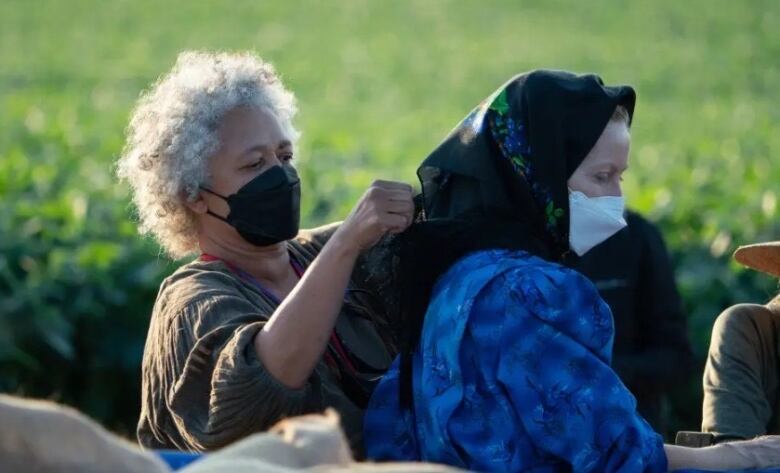 A woman adjusts the head scarf of another woman, who is dressed in traditional Mennonite clothing.