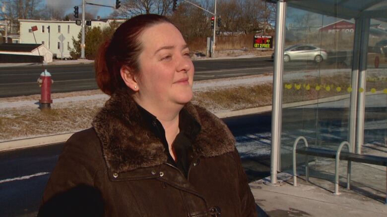 A woman is seen from shoulders up in a brown winter coat looking off the right side of the image, she is standing in front of a bus stop. 