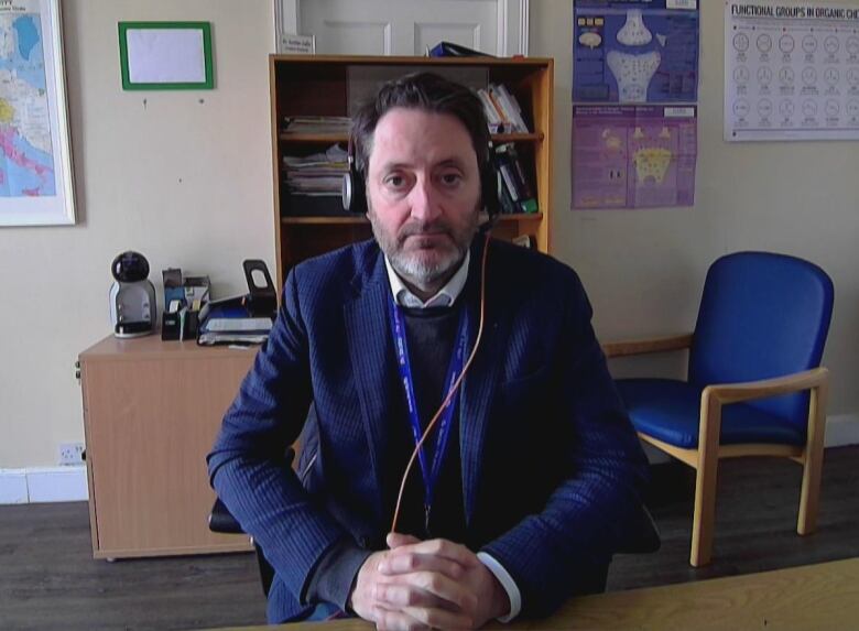 A person wearing headphones sits at a desk with their hands folded. Behind them is a shelf, a map of Europe and a diagram of a bone joint.