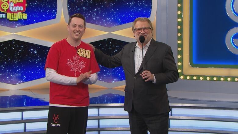 Two men stand together on The Price is Right stage, one with a microphone, placing his hand on the other's shoulder.