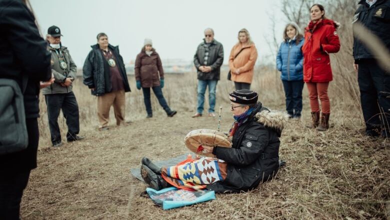 A woman sits on the ground drumming as other people stand around her in a circle.