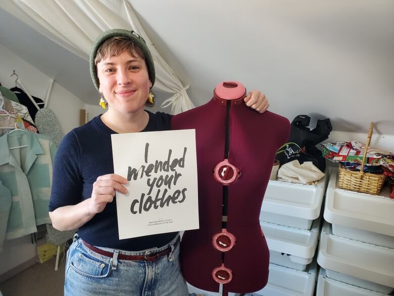 A smiling woman holds up a sign that says 'I mended your clothes,' while standing in her sewing room. 