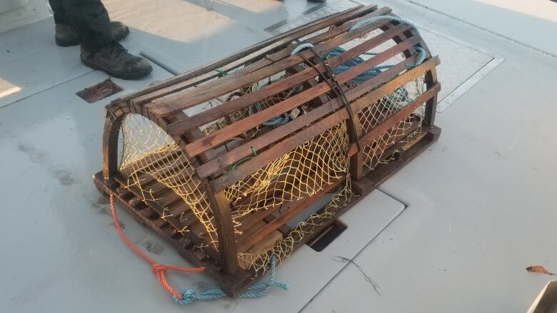 A brown lobster trap is seen on the grey deck of a boat.