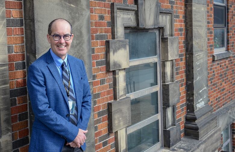 A man in a blue blazer and blue shirt with a striped blue tie stands with a red brick wall behind him.