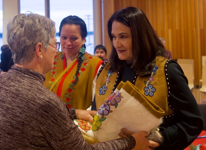 Two women in beaded hide vests stand side-by-side as another woman faces them, inside a room with people in the background.