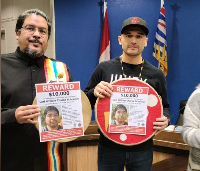 Two men are holding up reward posters with a picture of a man on each one. They are framed by a flag of B.C. and a flag of Canada at the local police station.