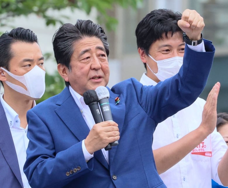 A man in a blazer speaks, gesturing with one hand while holding a microphone in the other hand.