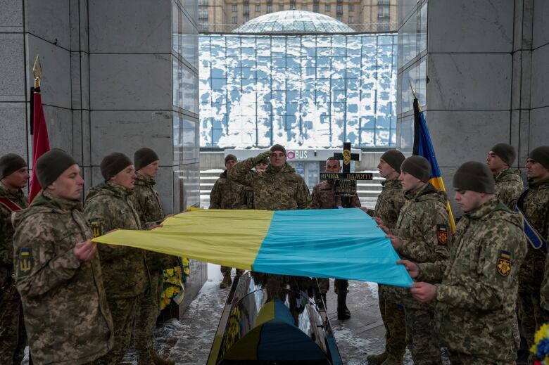 Several soldiers hold a Ukraine flag over a coffin.