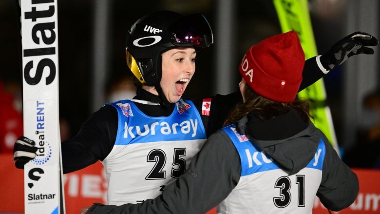 A woman wearing a helmet and holding skis opens her mouth and embraces a teammate.