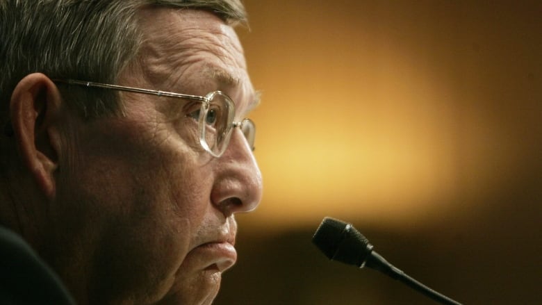 A closeup photo shows a side profile of a man with graying short hair, wearing glasses and frowning as he faces a microphone. 