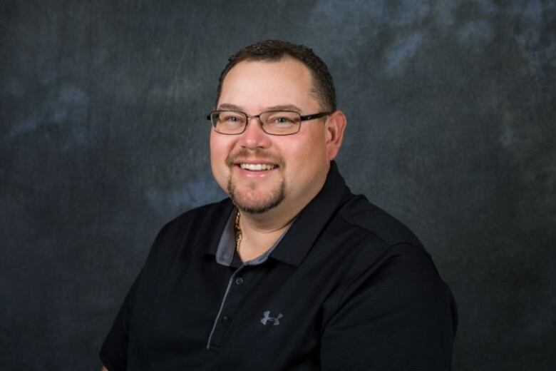 A man wearing a dark collared shirt and glasses smiles at the camera.