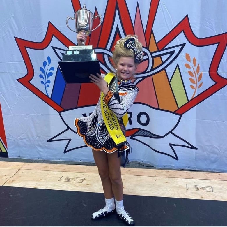 A young girl with a blonde wig holds up a trophy while wearing a sash and an elaborate costume.