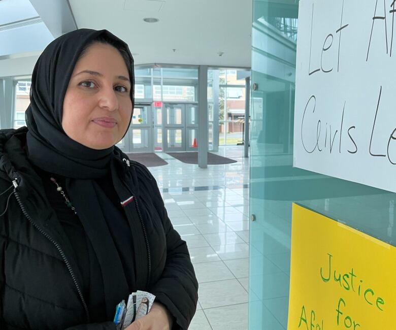 A middle-aged woman looks at the camera. She wears a hijab. She stands next to posters demanding justice for Afghan women and girls.