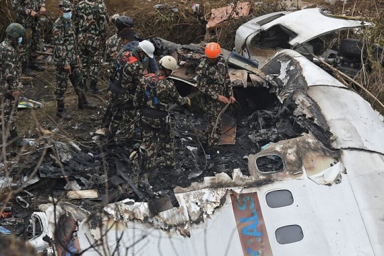 Uniformed rescuers in helmets look at part of a broken plane fuselage, which is lying on the ground in a forest.