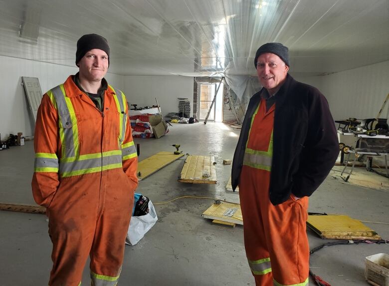 Two men wearing orange safety suits stand in a large, white, indoor space, which is currently under construction. 