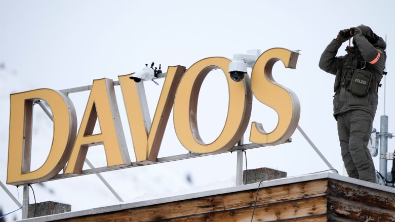 A police officer clad in grey holds up binoculars to his eyes as he stands beside a sign that says Davos.