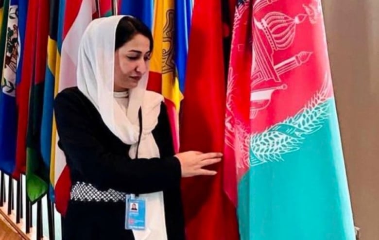 A woman in a black dress stands next to a row of national flags, gently touching the Afghanistan one.