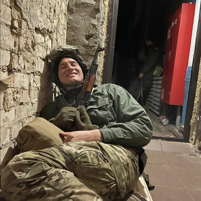 A man smiles, crouched in an alleyway. He's partially lying down, holding a gun and wearing a helmet.