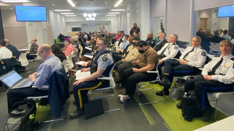 A crowd of residents, including those who are white and people of colour, sit alongside RCMP and Halifax Regional Police members on white chairs in a large carpeted room.