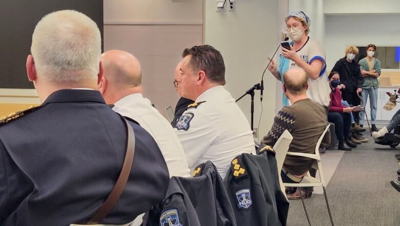 A white non-binary person with blonde hair and glasses stands at a microphone with their finger in the air as they look at a row of police officers.