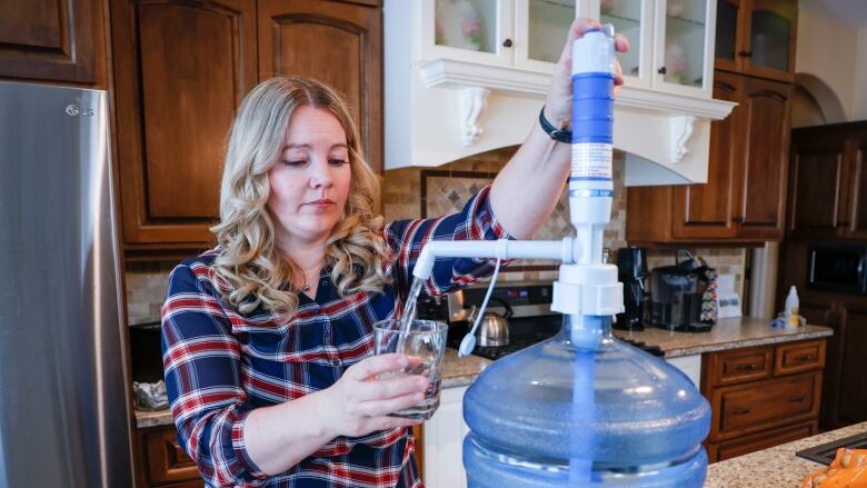 A woman in a plaid shirt pumps water from a reusable jug. She stands in a kitchen with brown cabinets.