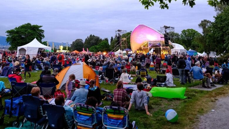 Concert goers sit on the grass at dusk in the summer. A stage with musicians is pictured in the background.