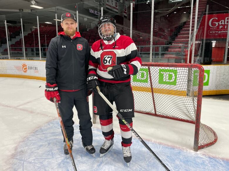 A coach and a player stand in front of a hockey net on an ice rink.