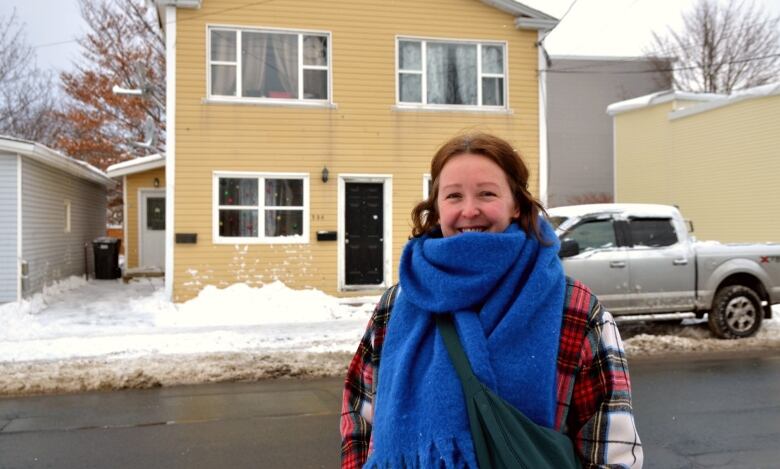 A woman with a blue scarf and red coat stands in front of a yellow house. To her right is a silver truck. 