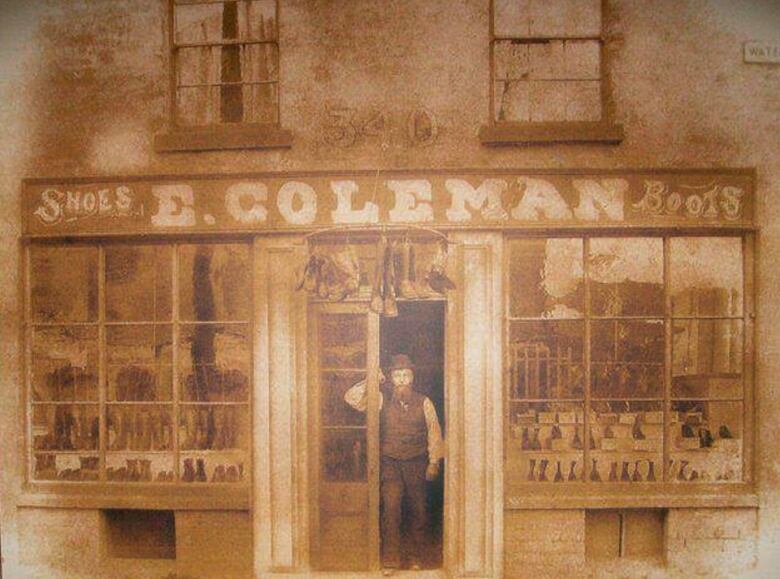 A faded photo from the 1800s of a store front. A man stands in the doorway with a sign above that reads E. Coleman. 