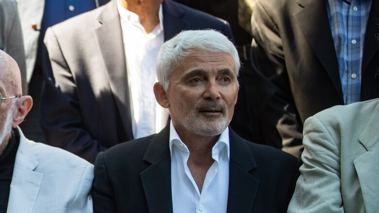 A white-haired man in a suit sits with his hands folded.