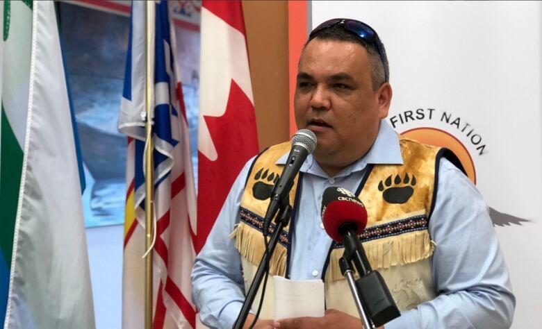 A man speaks into a microphone, standing in front of flagpoles.