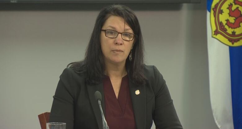 A woman sits at a desk with a microphone and a Nova Scotia flag behind her.