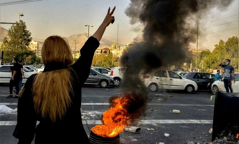 A woman with her back to the camera makes a 'V for victory' gesture near a burning tire.