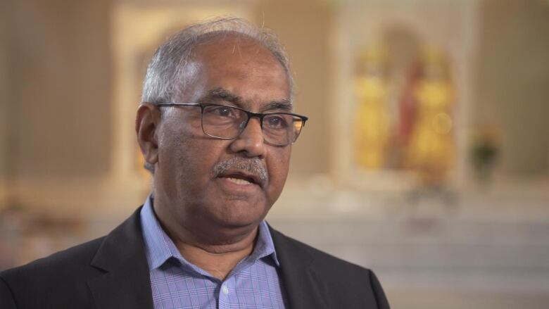 A man with glasses, grey hair and a short mustache is speaking to behind the camera. The background behind him is blurred.