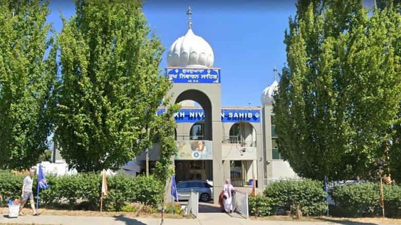 A grey building with pillars, a white dome, and white block text against a blue block background is pictured surrounded by trees.