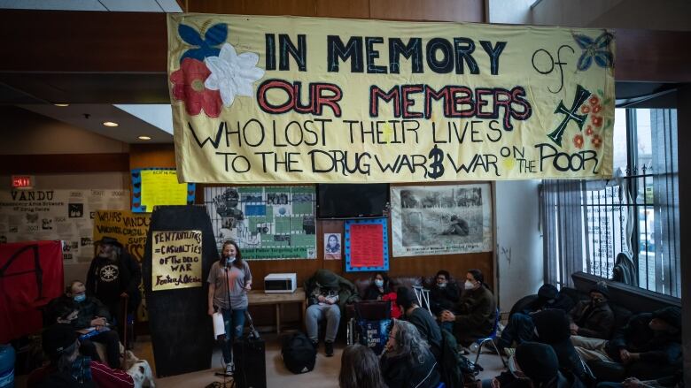 A woman speaks at a mic in front of a group of people in a small room. A banner hangs above them reading 'In memory of our members who lost their lives to the drug war and war on the poor'.