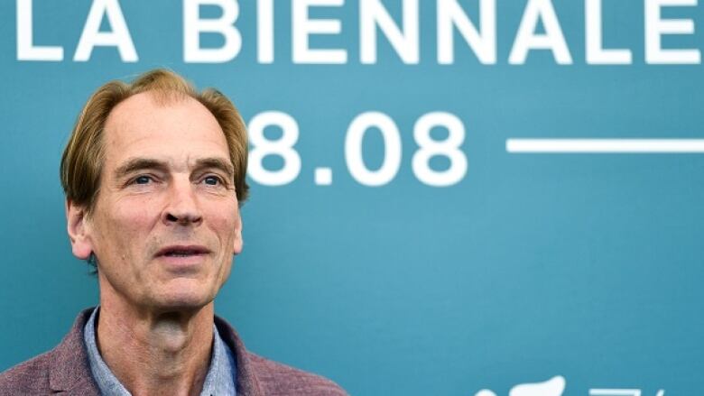 The actor Julian Sands appears in front of a teal blue backdrop at a film festival.