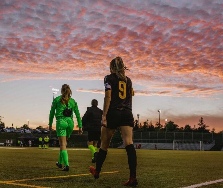 Two women walk onto a soccer field with a pink sunset above. 