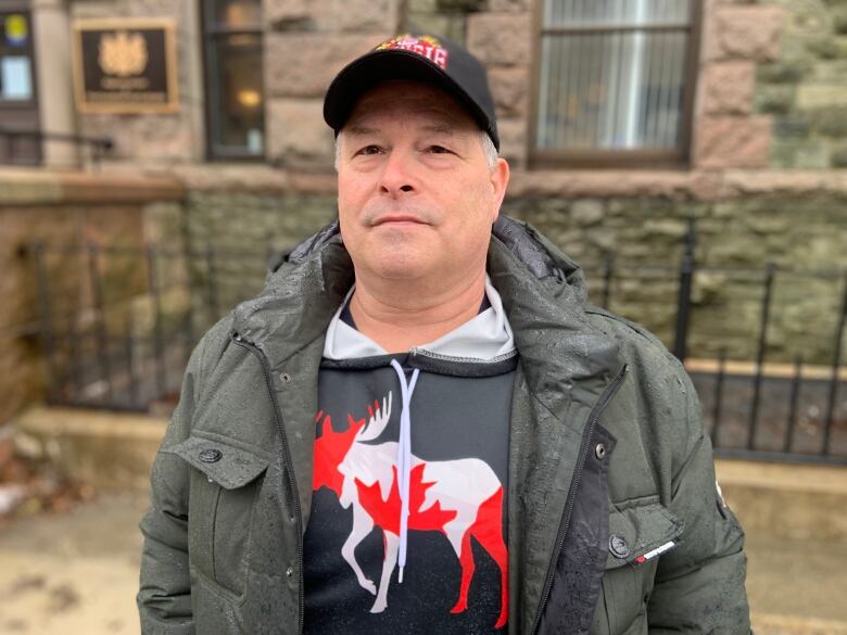 A person stands outside a stone building. They're wearing a hoodie featuring an illustration of a moose with the colours of the Canadian flag.