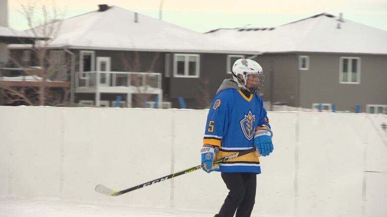 A hockey player wearing a team jersey, helmet and gloves holds his stick while skating on an outdoor rink. 