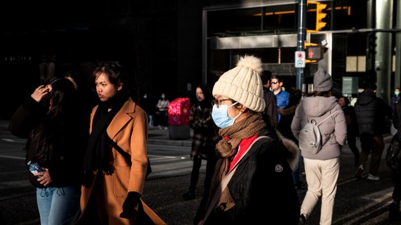 A crowd of people walk through downtown Vancouver. In the foreground is a woman with a beige tuque and brown scarf, wearing a blue surgical mask.