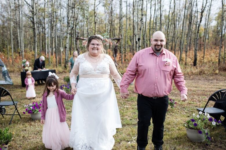 A woman in a flowing white dress holds hands with a man in a pink shirt, and a girl in a pink dress, in a forest environment.