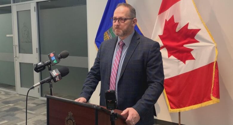 A middle-aged white man stands a podium before flags for Lethbridge and Canada. 