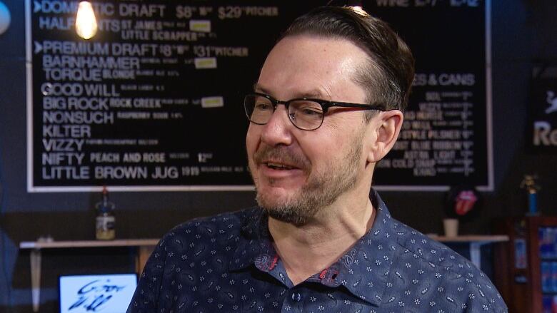 A man wearing a navy button-down shirt and glasses speaks to someone off-camera.