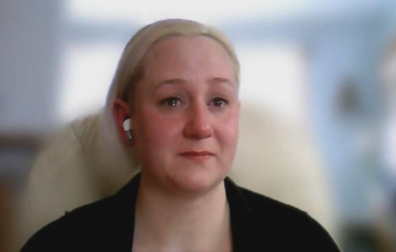 A blonde woman sits with a black jacket and watery eyes