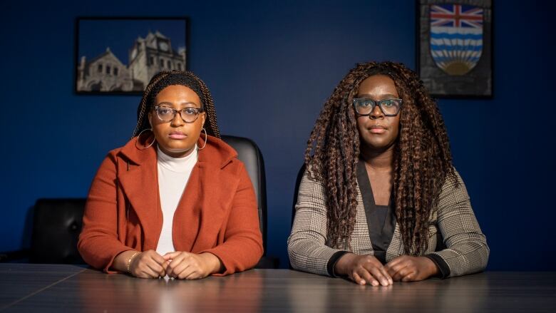 Two Black women are shown sitting at a table in a conference room directly facing the camera, in front of a blue wall. The woman on the left is wearing a burnt orange jacket, white turtleneck and hoop earrings. The woman on the right has thick-rimmed black glasses and a grey plaid dress.