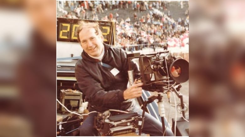 A man holds camera equipment in a stadium, wearing a black jacket and smiling.